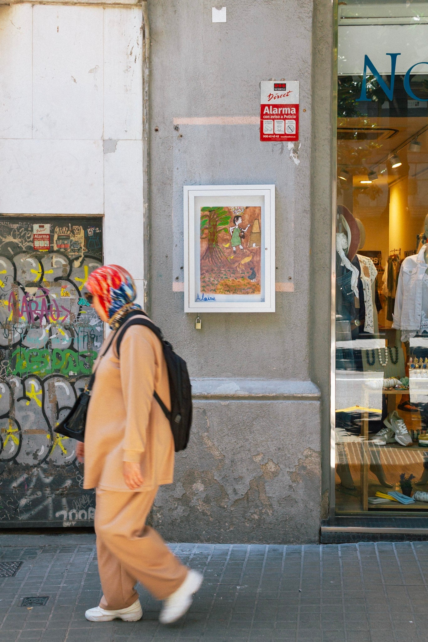 Street-level view of La Finestra, the public window gallery of Aloïse at Gran de Gràcia 123, showing a flat, unframed artwork on display inside the exterior glass vitrine.
